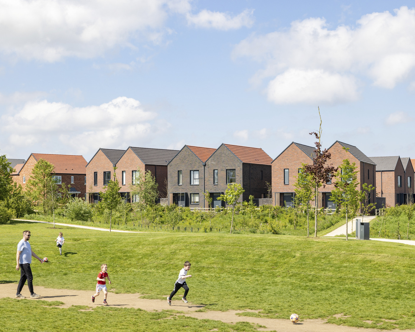 homes in the background with people walking through a green park on a sunny summer day, trees in full leaf and a bright blue sky overhead.